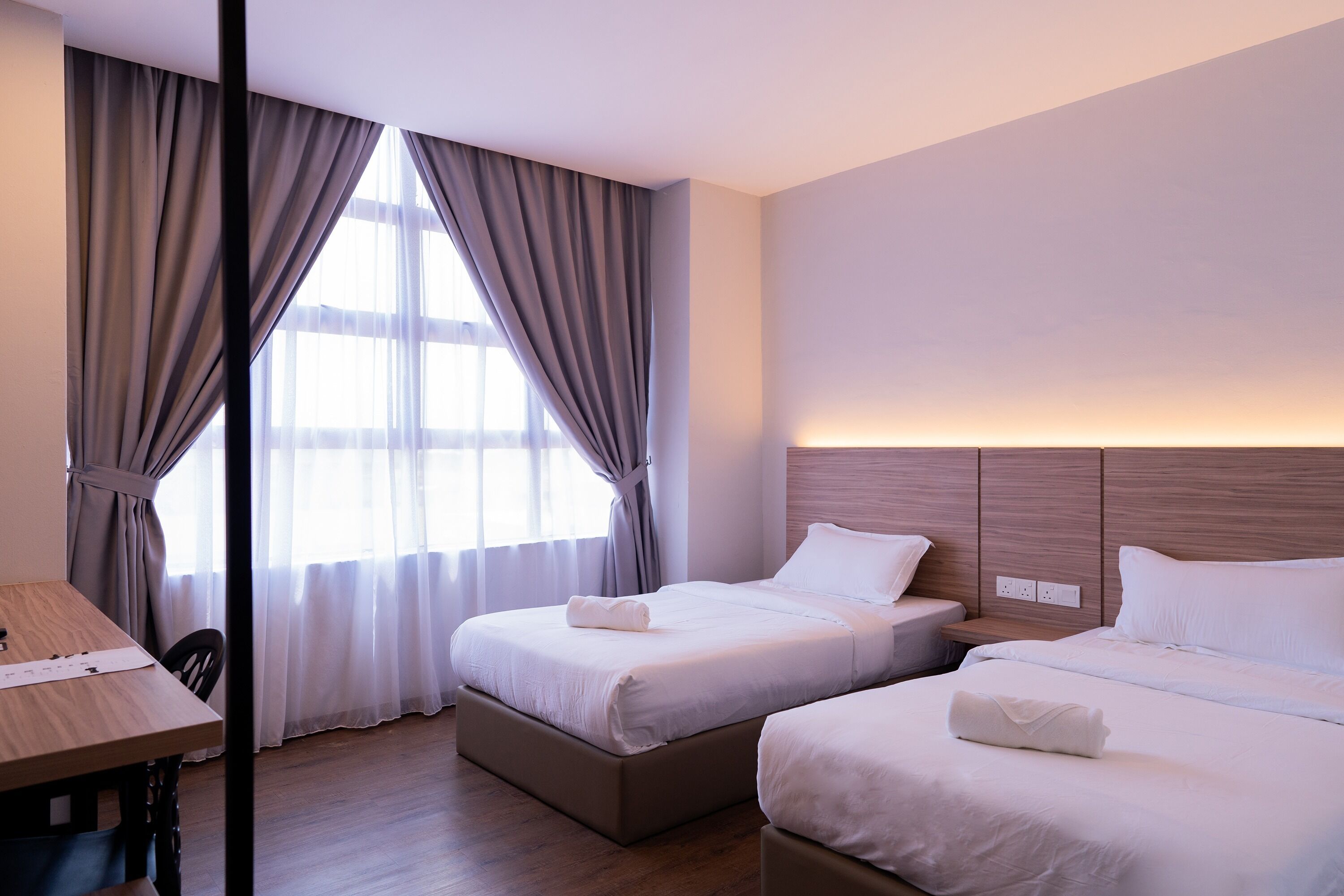 A hotel room with two beds, a wooden desk, and a view of the outdoors through large windows.