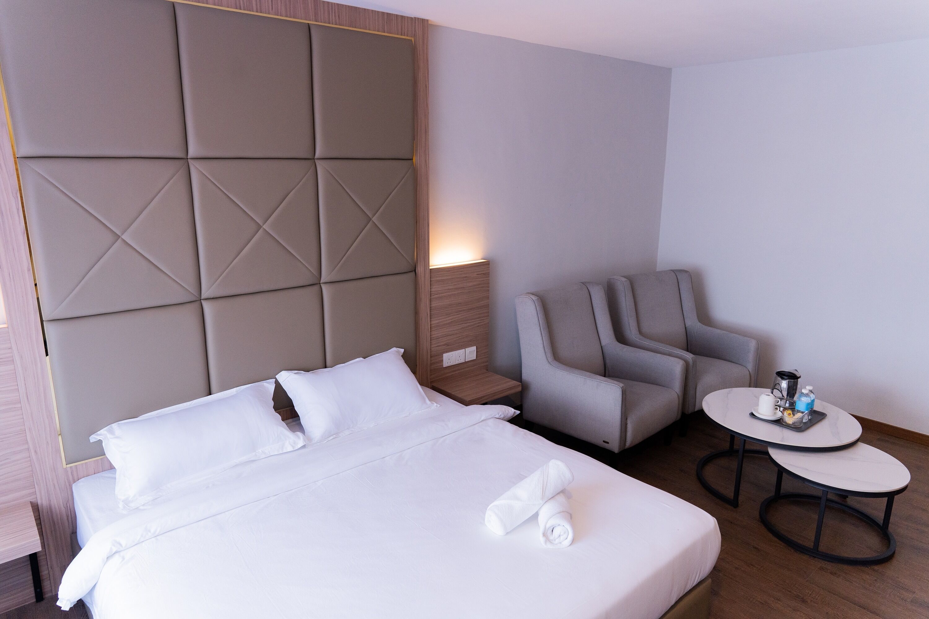 A hotel room with a large bed, a seating area with a chair and a small table, and a wall panel with a light.