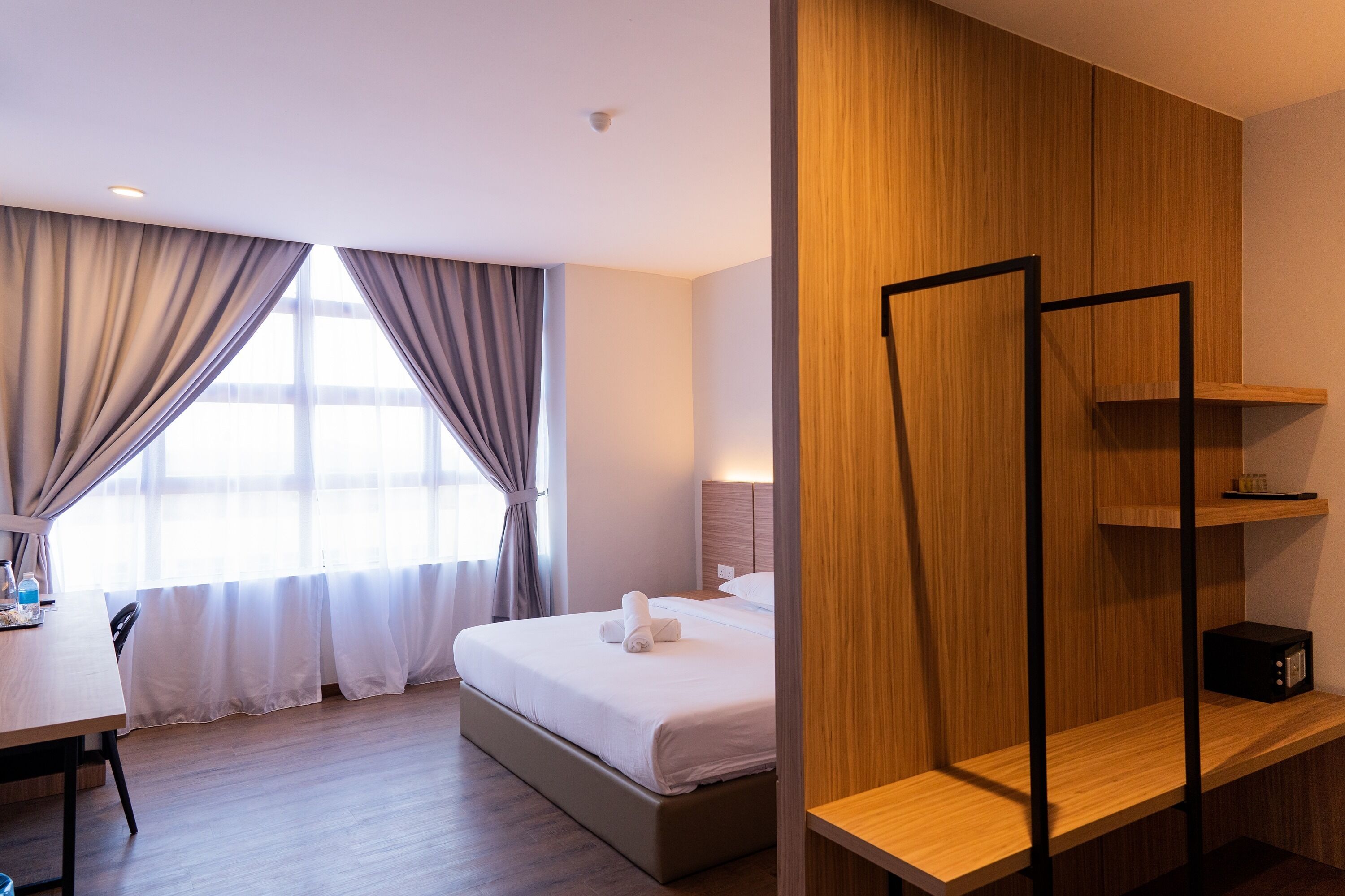 A hotel room with a bed, a desk with a chair, a bedside table, and a wardrobe.