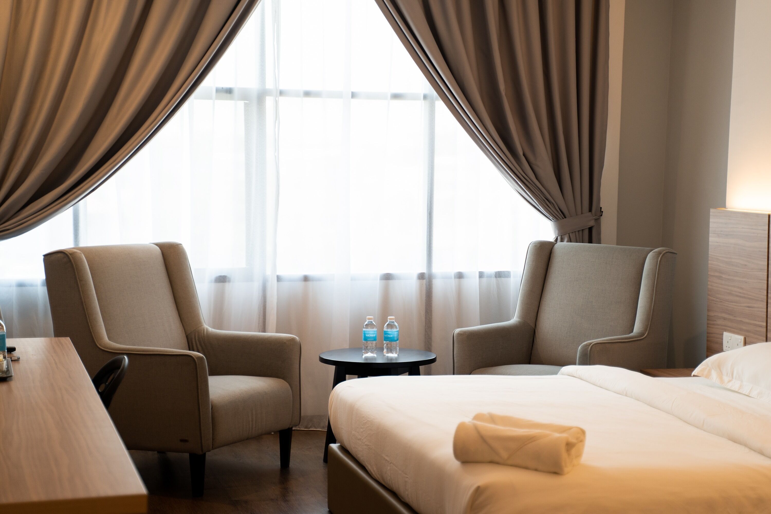 Habitación de hotel con cama, dos sillones, una mesita con botellas de agua y vistas al exterior a través de cortinas traslúcidas.