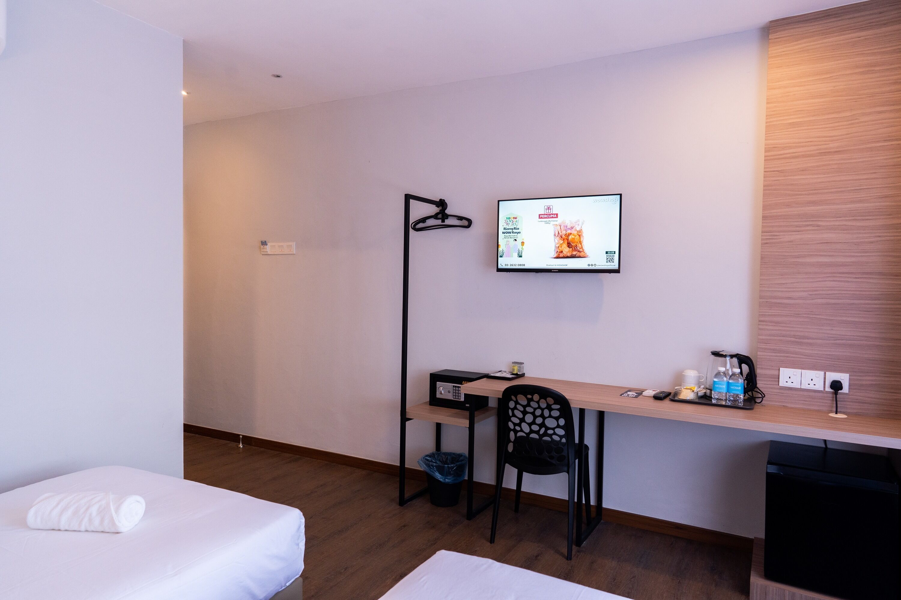 A hotel room with a bed, a desk with a computer, a chair, a TV mounted on the wall, and a small trash bin.