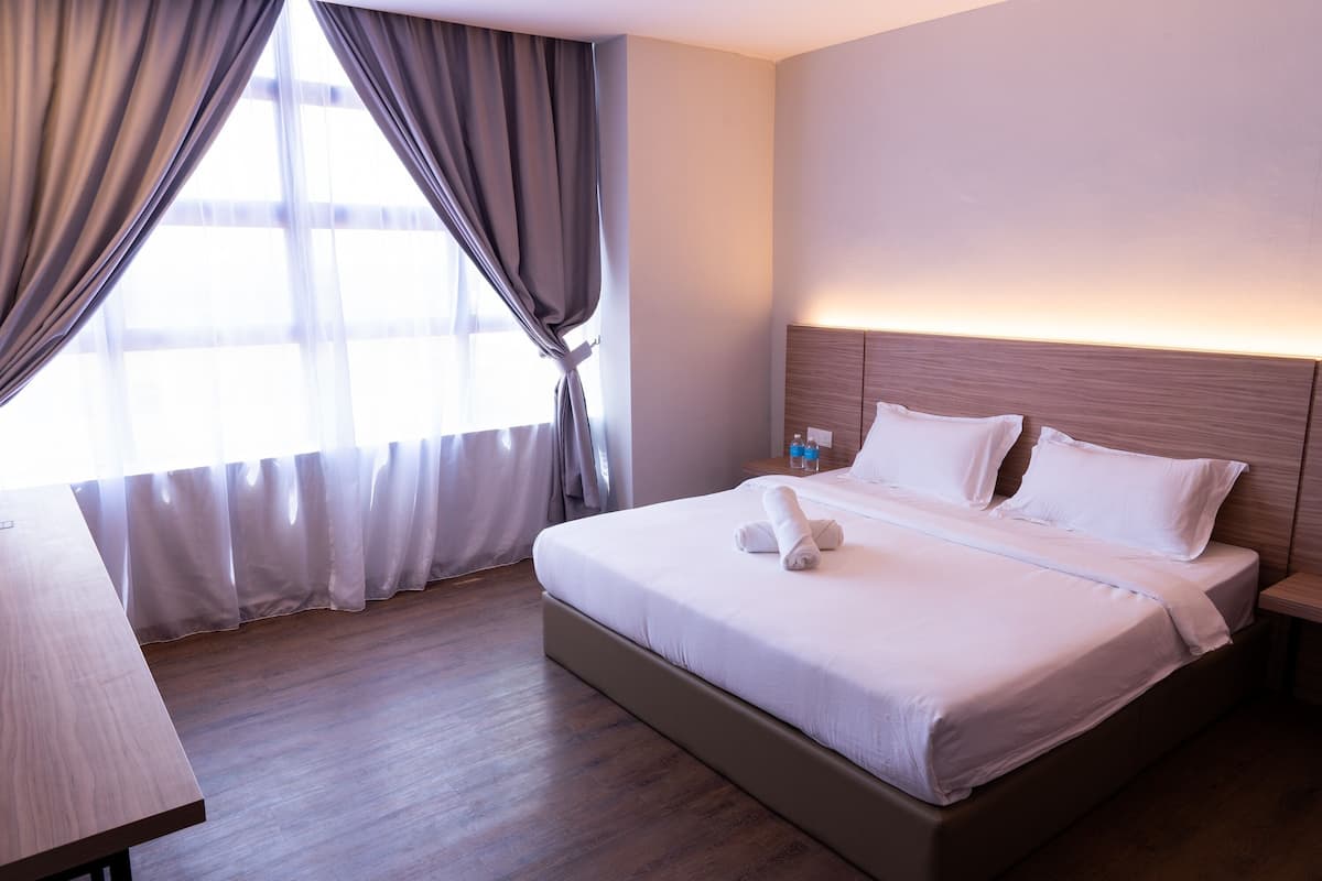 Ein Hotelzimmer mit einem großen Bett, einem Nachttisch aus Holz, einem Schreibtisch und einem Fenster mit Vorhängen.