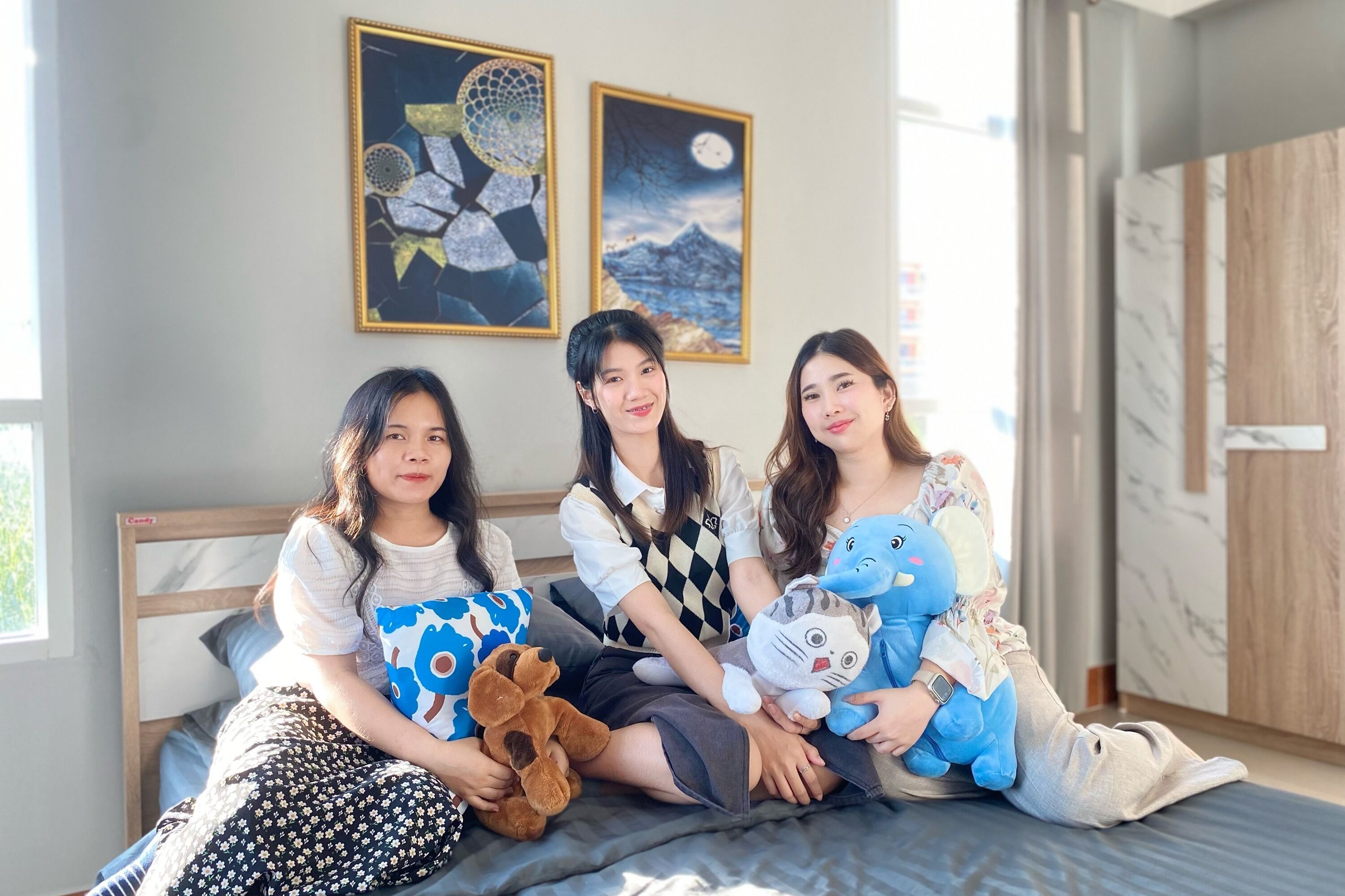 Three people sitting on a bed in a room with a wooden headboard, two framed pictures on the wall, and a window with daylight coming through.