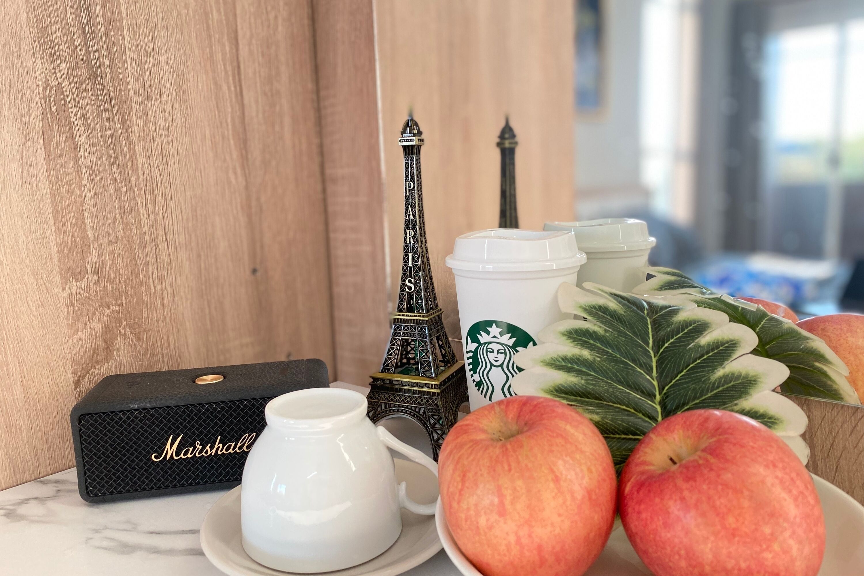 A table with a white teapot, a black box labeled "Marshall," two Eiffel Tower figurines, a Starbucks cup, and a bowl of apples.