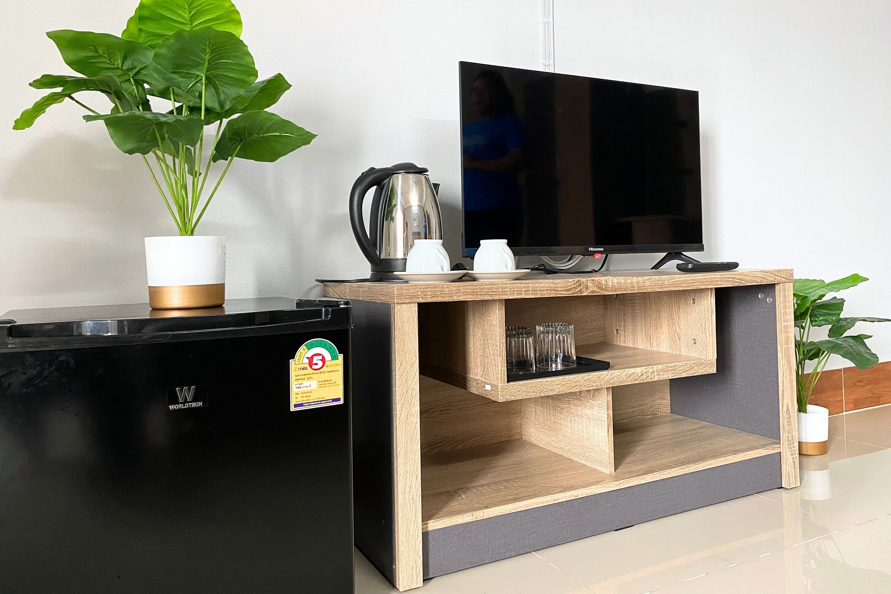 A compact kitchenette with a black refrigerator, a wooden TV stand, a kettle, and a potted plant.