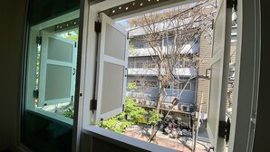 A view from a window with a white frame, looking out to a balcony area with a metal railing, a tree, and a building in the background. - Forgotten Hostel Silom (Bangkok)