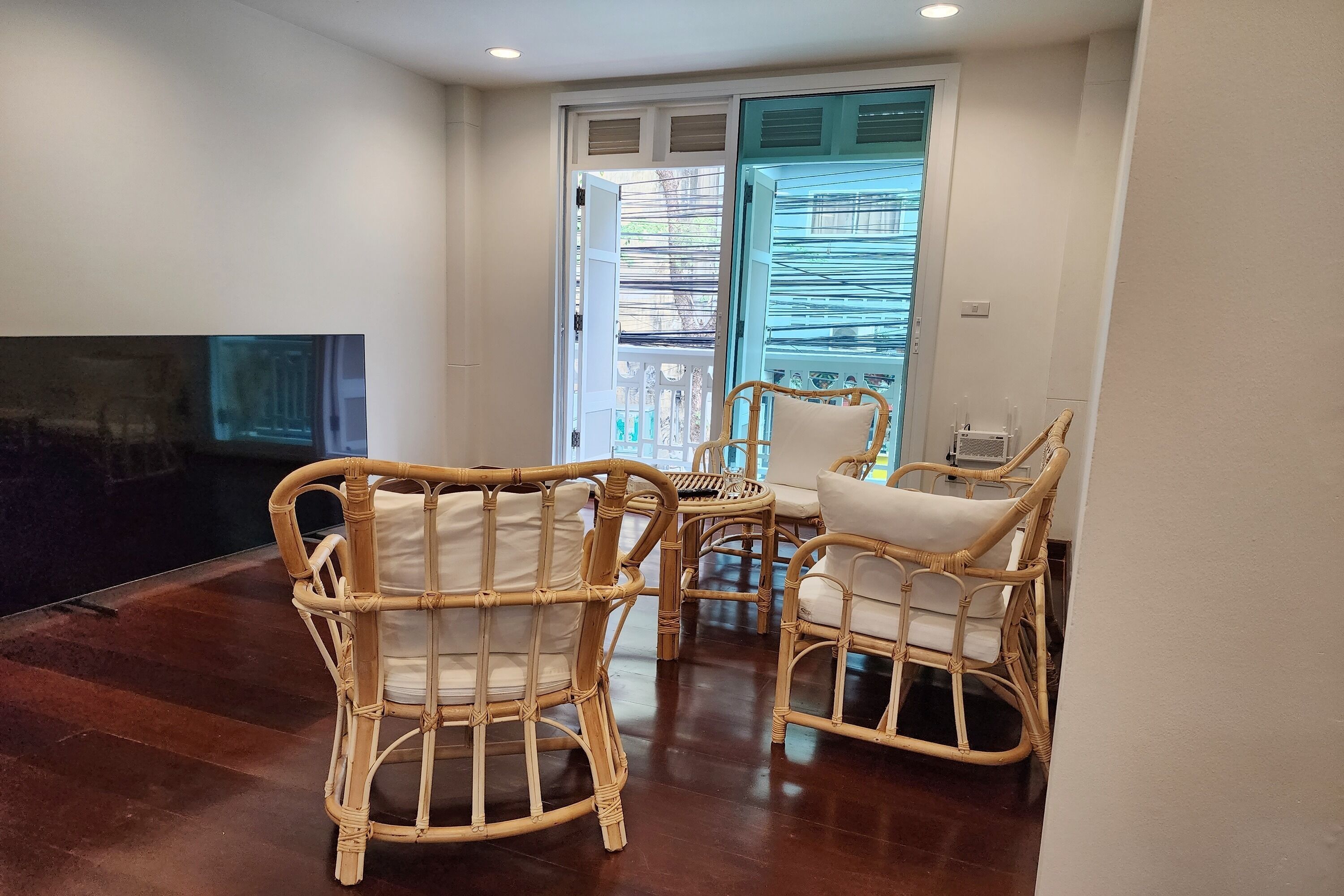 Una habitación con piso de madera, dos sillas de mimbre con cojines, una mesa pequeña y un televisor a la izquierda.