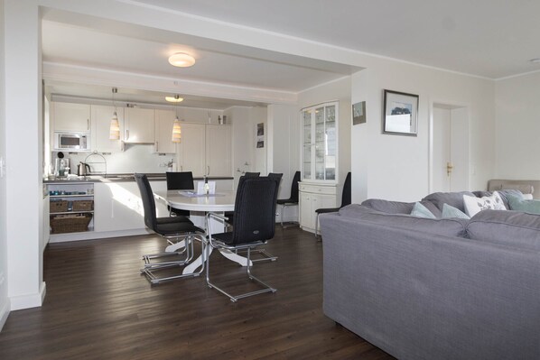 A modern living room with a grey sofa, a dining table with chairs, and a kitchen area with white cabinets and a microwave.