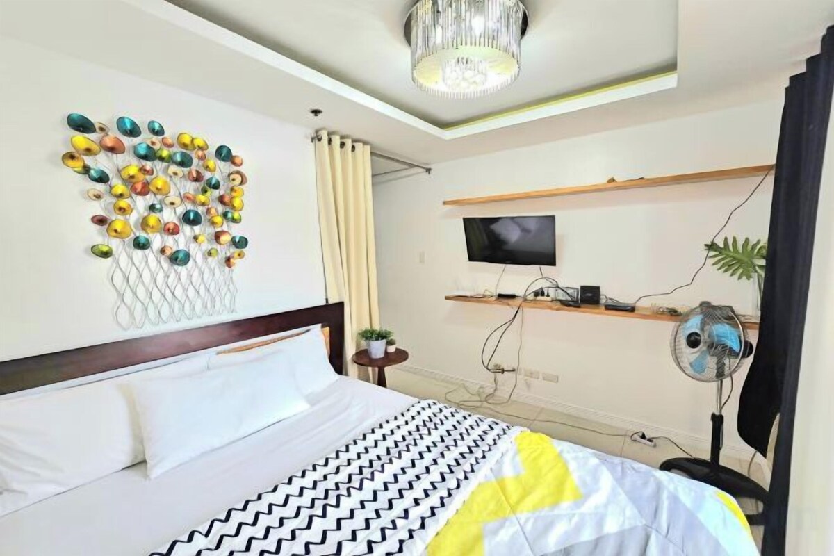 A hotel room with a bed, a wall-mounted TV, a ceiling fan, and a decorative wall art piece.