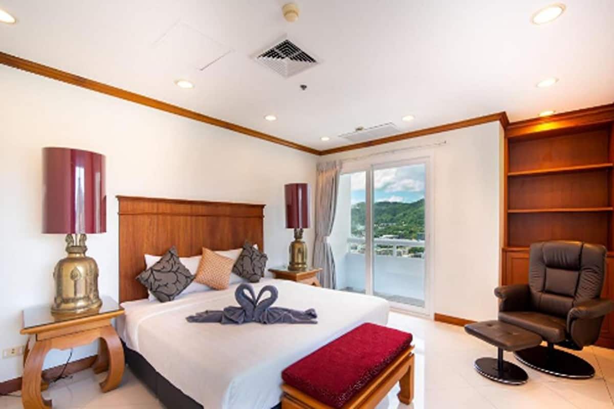A hotel room with a large bed, two bedside tables with lamps, a red throw blanket, a leather armchair, and a view of the outdoors.