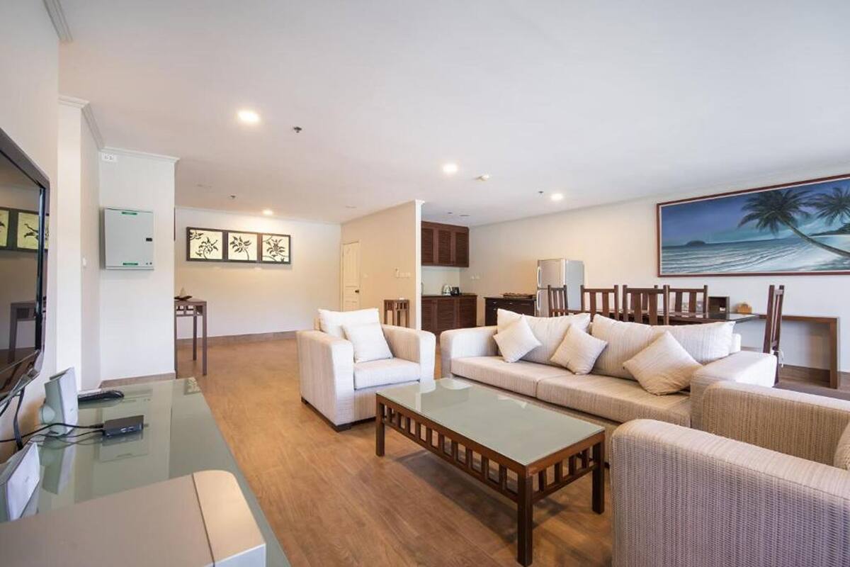 A spacious living area with a sofa set, a coffee table, and a dining table. There is a kitchen area in the background with cabinets and a refrigerator.