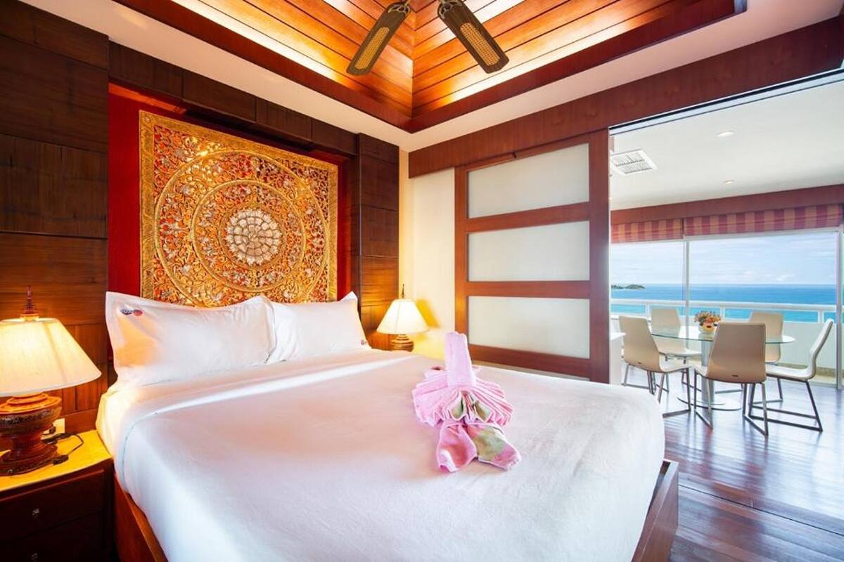A hotel room with a large bed, a decorative headboard, a view of the ocean, and a balcony with outdoor seating.