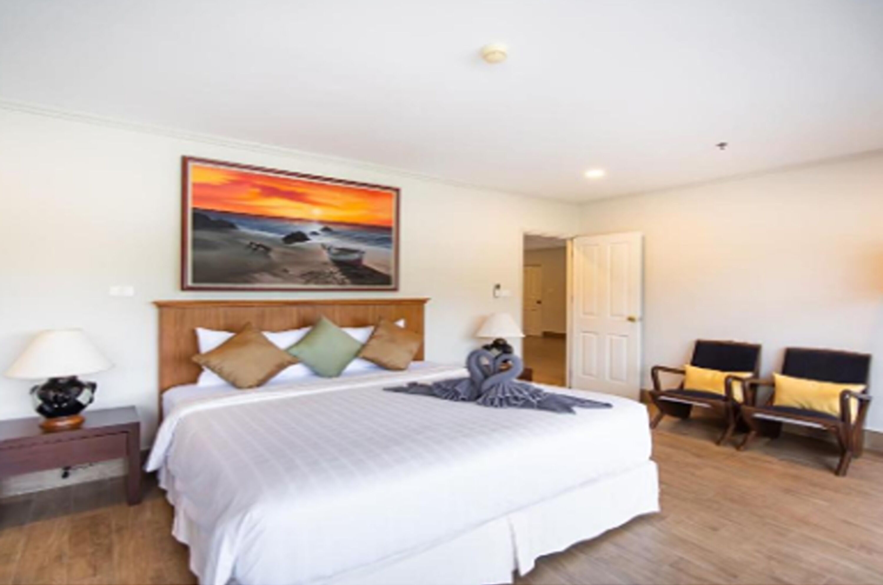 A hotel room with a large bed, a painting of a sunset, a wooden headboard, two bedside tables with lamps, and a door leading to another room.