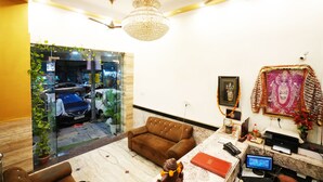 A well-lit room with a chandelier, a brown sofa, a desk with a computer, and a glass wall with a view of the street. - Hotel Le Sancy (New Delhi)