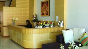 A reception area with a curved wooden reception desk, a brick wall, and a large decorative painting above it. There are two people seated at the desk, and a sofa with colorful cushions in the foreground. - Fruit Paradise Hotel (Patong)