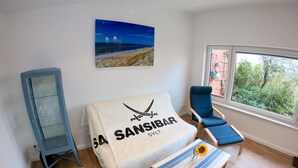 Un soggiorno moderno con un quadro a tema spiaggia, un divano con un asciugamano, una poltrona blu e un tavolino con un girasole.