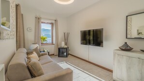 A living room with a sofa, a TV, a cabinet, a window with a view, and a decorative mirror. - Casa Magnolia Varenna (Varenna)