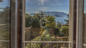 A view through a window with a balcony, overlooking a body of water, mountains, and a garden. - Casa Magnolia Varenna (Varenna)