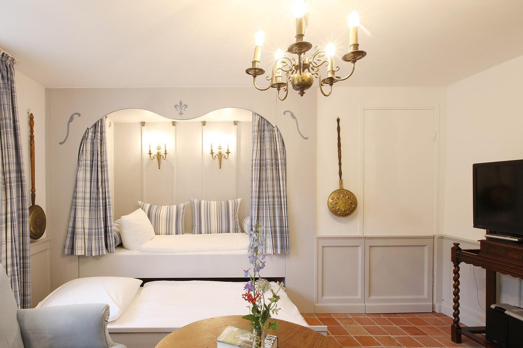A hotel room with a bed, a television, a chandelier, and a mirror with curtains.