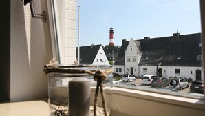 A view from a window with a jar in the foreground, a white building with a red and white lighthouse in the background, and parked cars on the street. - Top renovated apartment ideal for 3 persons (Hörnum auf Sylt)