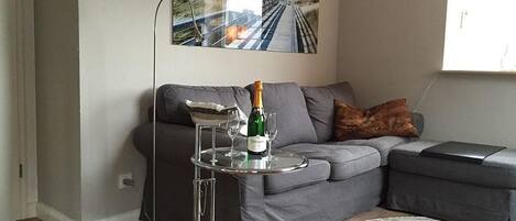Una sala de estar moderna con un sofá gris, una mesita redonda con una botella y vasos, una lámpara de pie y un cuadro abstracto grande en la pared.