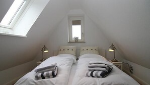 Una cama doble con ropa de cama y toallas blancas, dos lámparas de noche, un tragaluz y una pequeña ventana sobre la cama.