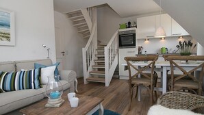 Una sala de estar moderna con una mesa de centro de madera, un sofá con cojines a rayas y una escalera que lleva a una zona de cocina con armarios blancos y una mesa de comedor.