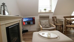A cozy living room with a fireplace, a television displaying 'NATIONAL OFFICE', and a dining area with wooden chairs and a table. - For a family of 4 or for two couples.... (Hörnum auf Sylt)