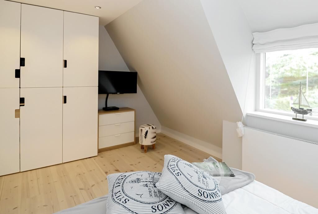 Quarto com teto inclinado, cama com almofadas estampadas, televisão sobre um suporte, um banquinho pequeno e um guarda-roupa.