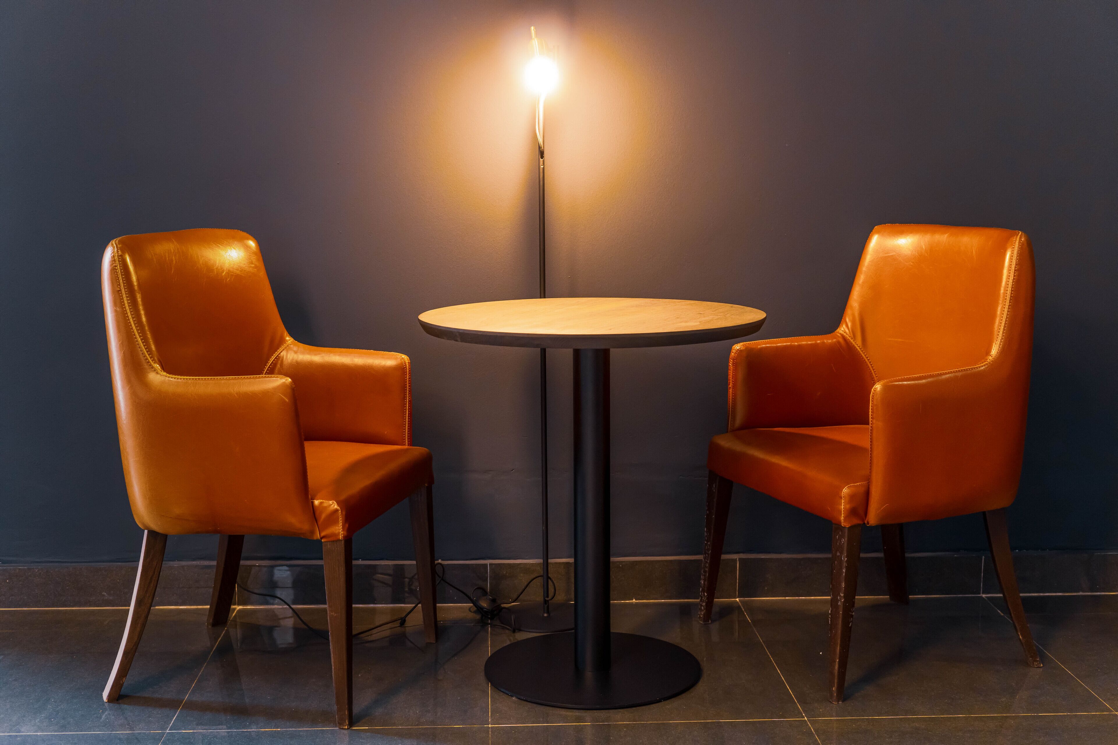 A small round table with two leather chairs, a floor lamp, and a dark wall.