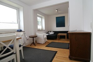 Una habitación con piso de madera, paredes blancas, una silla blanca, un sofá blanco, una mesita de madera y un tapete oscuro.