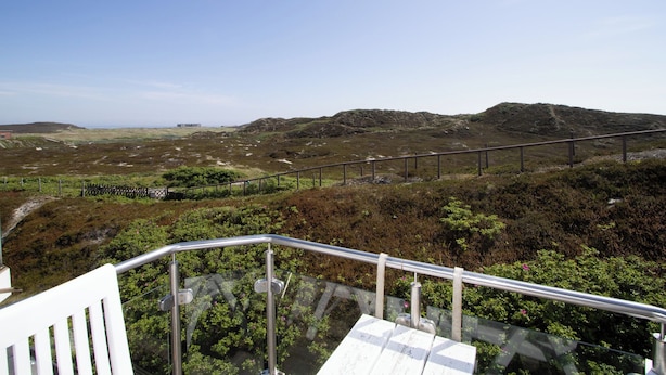 Vista panorâmica de uma varanda com guarda-corpo metálico, com vista para uma paisagem acidentada e vegetação rasteira, e um edifício ao longe.