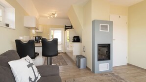 Ein modernes Wohnzimmer mit einem grauen Sofa, schwarzen Sesseln und einem weißen Kissen mit Anker-Muster. Im Hintergrund ist eine Küche mit weißen Schränken und einer schwarzen Kaffeemaschine zu sehen.