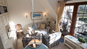 Ein gemütliches Wohnzimmer mit Sofa, Sesseln und Esstisch. Ein Fenster mit Vorhängen, eine Heizung und eine Wandlampe sind ebenfalls vorhanden.