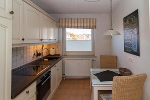 Une cuisine avec des meubles blancs, un pare-feu en carrelage, une fenêtre avec des stores, un luminaire suspendu et un tableau noir servant de menu, fixé au mur.