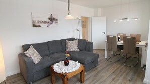A modern living room with a grey sofa, a wooden coffee table with a vase, and a dining area with a white table and brown chairs. - Fantastic vacation apartment in Hörnum for 4 people - pure relaxation! (Hörnum auf Sylt)