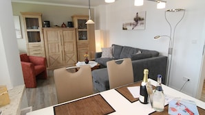 A modern living room with a grey sofa, a red armchair, and a dining table with a bottle of champagne. There are cabinets in the background and a floor lamp. - Fantastic vacation apartment in Hörnum for 4 people - pure relaxation! (Hörnum auf Sylt)