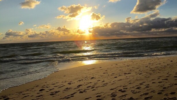 Ein Strand mit Fußspuren, die zum Wasser führen, ein Sonnenuntergang im Hintergrund und ein klarer Himmel.