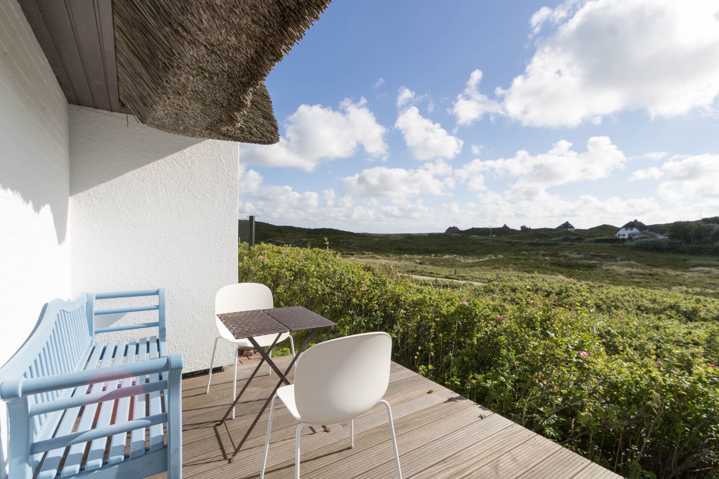 Une terrasse en bois surmontée d’un toit de chaume, avec une chaise blanche, une chaise bleue et une petite table.
