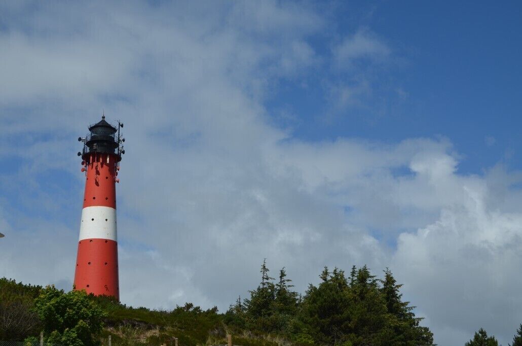 Un faro con torre a rayas rojas y blancas, rodeado de vegetación y un cielo parcialmente nublado.