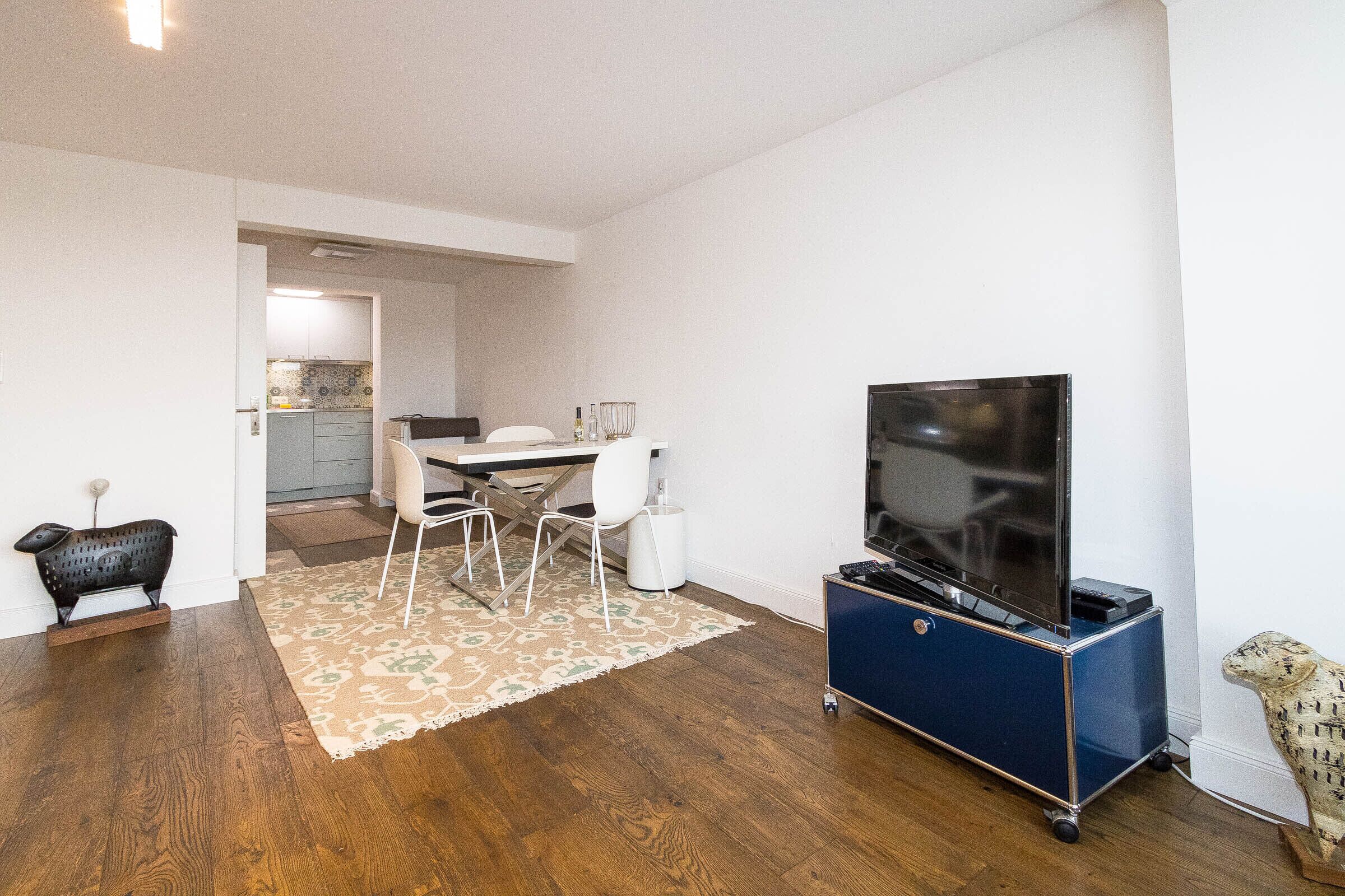 Un salon moderne avec un téléviseur à écran plat posé sur un meuble, un coin repas avec une table et des chaises, et une cuisine en arrière-plan.