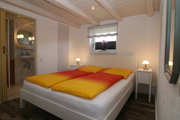 Une chambre à coucher avec un lit, deux lampes de chevet, une fenêtre avec des stores et une salle de bain visible à travers une porte ouverte.