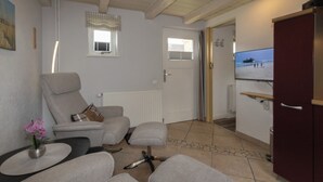 En stue med sofa, lænestol og skammel, et lille bord med en vase, et fjernsyn og en dør der fører til et andet rum.