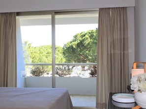 Ein Hotelzimmer mit großem Fenster, einem Bett, einem Nachttisch mit Lampe und einer Vase mit Blumen.