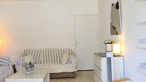 Une petite pièce, peu meublée, avec un canapé à rayures, une petite table surmontée d’un récipient en verre, une commode blanche et un radiateur.