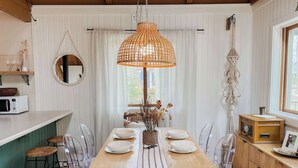 Ein Essbereich mit einem Holztisch, klaren Stühlen, einer hängenden orangen Lampe und Blick ins Freie durch ein Fenster.