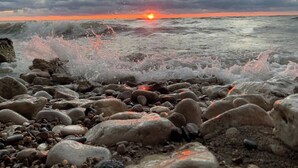 Eine felsige Küstenlinie, auf die Wellen auf die Kiesel treffen, mit einem Sonnenuntergang im Hintergrund.