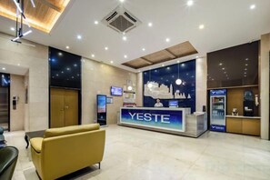Un espace d’accueil comprenant un comptoir de réception portant l’inscription « YESTE », un coin salon avec un canapé et une grande fenêtre offrant une vue sur le paysage urbain.