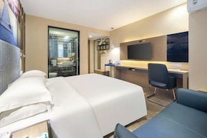 Quarto de hotel com uma cama, uma escrivaninha, uma cadeira, uma televisão e um banheiro visível por uma porta de vidro.