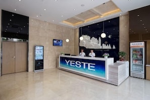A reception desk with a sign reading 'YESTE' in a modern lobby with a cityscape mural, a digital display, and a beverage refrigerator. - Yeste Hotel (Tianyang) (Baise)